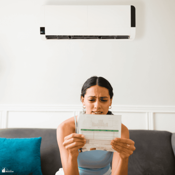 Person sitting beneath a wall-mounted air conditioner while reviewing a high electricity bill, illustrating rising home energy costs and household budget concerns.