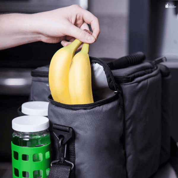 Black insulated lunch bag being packed with bananas beside reusable drink containers for carrying meals to work or school.