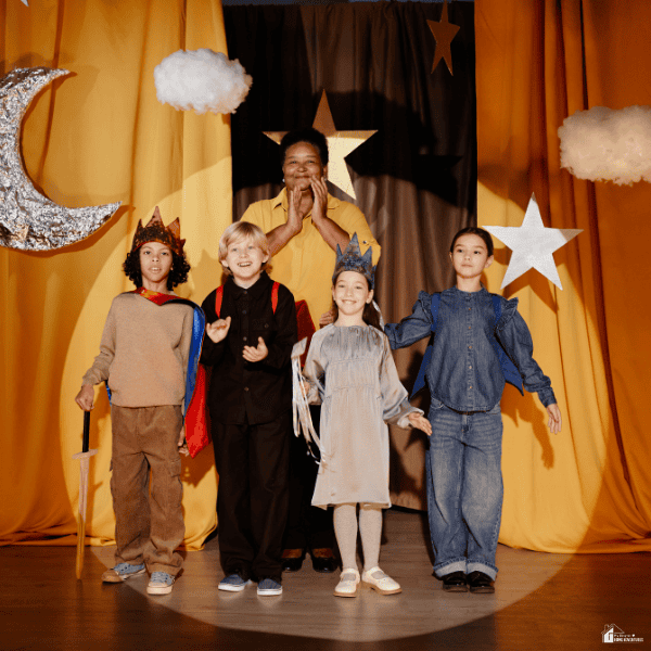 A group of young children in costumes stand together on a decorated school stage after participating in a classroom performance.