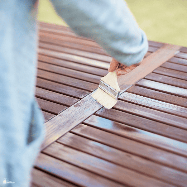 Person applying a protective coating to a wooden deck with a brush to help seal the surface against moisture and weather damage.