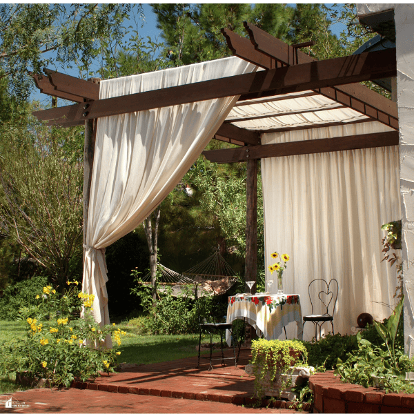 Outdoor patio with flowing white curtains around a wooden pergola, surrounded by lush greenery and a small dining area.