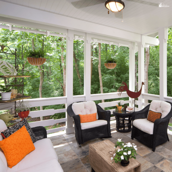 Covered patio with wicker seating, bright accent pillows, hanging planters, and an open view of a wooded backyard.