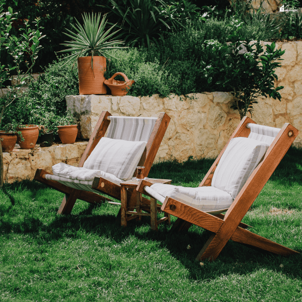 Low-upkeep backyard features include wooden lounge chairs, a grassy seating area, stone garden beds, potted plants, and established landscaping.