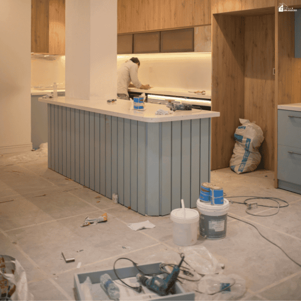 A partially renovated kitchen with an unfinished island, construction tools, paint supplies, and a worker completing updates as part of a budget home renovation.