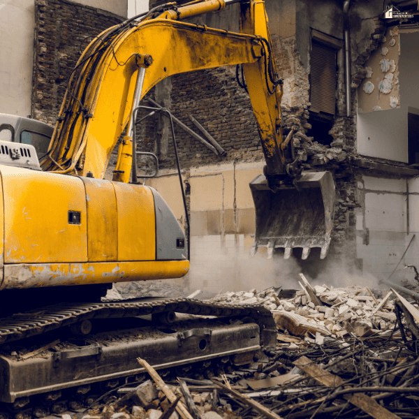 Large excavator tearing down a house wall with rubble and construction debris surrounding the demolition site.