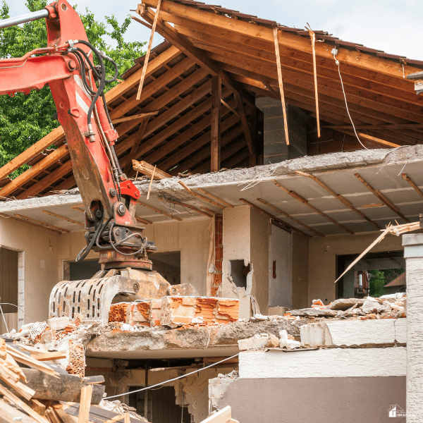 Excavator removing brick walls from a partially demolished house with exposed roof framing and debris around the property.