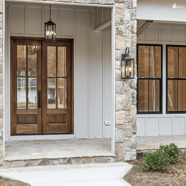 Wooden double entry doors with glass panels beside black-framed windows on a stone-accented home exterior, showing features homeowners may consider when planning replacements.