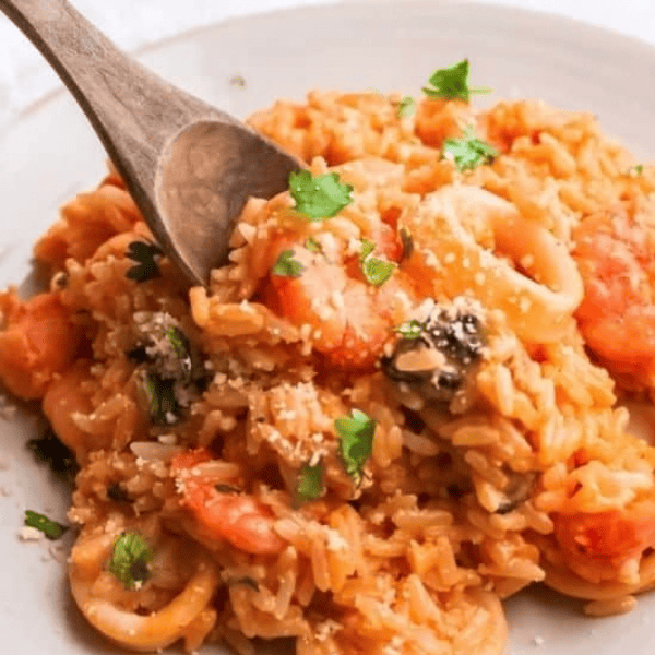 Arroz con Mariscos: A white bowl holds orange seafood rice with shrimp, squid rings, shellfish, and chopped herbs beside a wooden spoon.