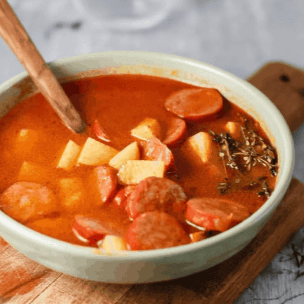 Chorizo and Potato Soup: A pale green bowl holds red broth with sliced chorizo and potato chunks beside a wooden spoon.