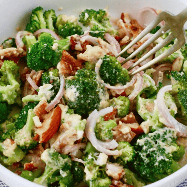 Easy Broccoli Salad with Bacon: Broccoli florets, bacon pieces, sliced red onion, and almonds fill a bowl with creamy dressing and a serving fork.