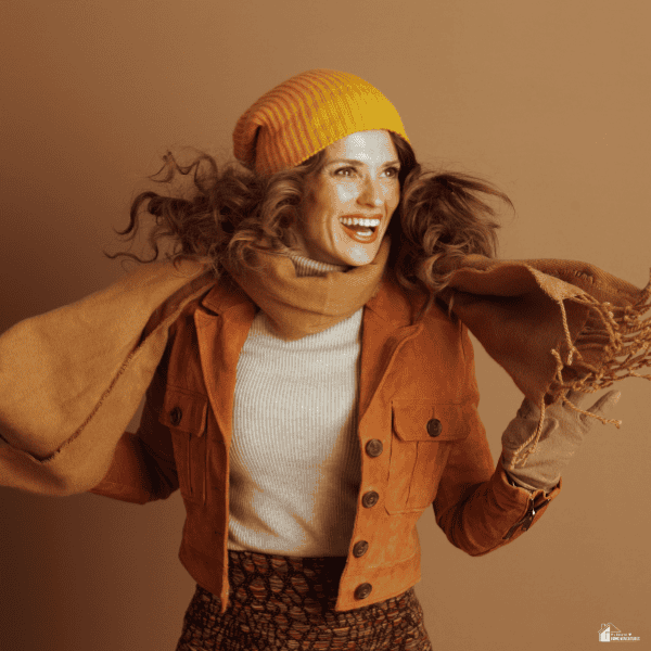 A woman models fall fashion essentials, pairing a rust jacket with a knit hat, scarf, and patterned skirt.