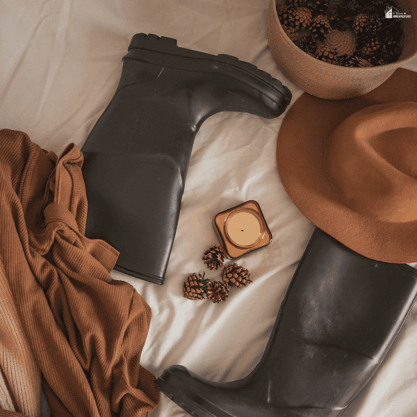 Black rain boots, a brown felt hat, and a rust-colored garment are arranged as fall fashion essentials.