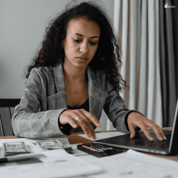 A woman uses a laptop beside a calculator, paperwork, and cash while managing her finances with a budgeting app.