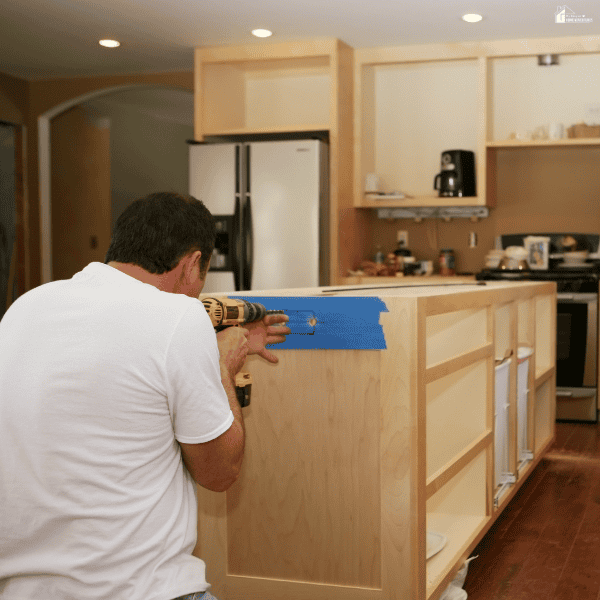 A man uses a power drill on an unfinished wooden island during a kitchen remodel, with cabinets and appliances behind him.