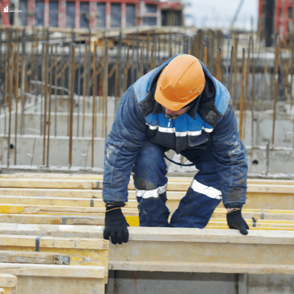 A worker in an orange hard hat positions wooden formwork beside exposed steel reinforcement.