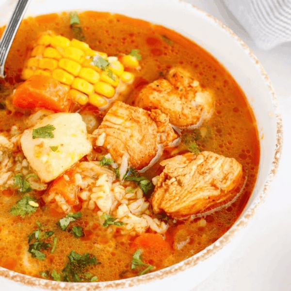 Puerto Rican Chicken Soup: Chicken pieces, corn, potatoes, and carrots fill a white bowl of golden broth garnished with chopped herbs.