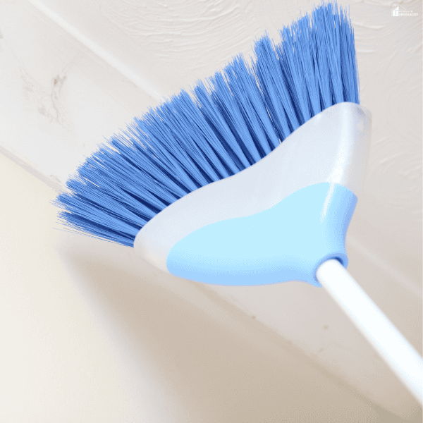 A long-handled broom removes webs from a ceiling corner to help prevent spiders from settling indoors.