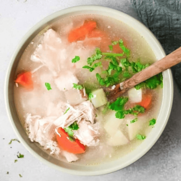 Sopa de Pollo: Shredded chicken, carrots, potatoes, and thin noodles fill a bowl of light broth beside a wooden spoon.