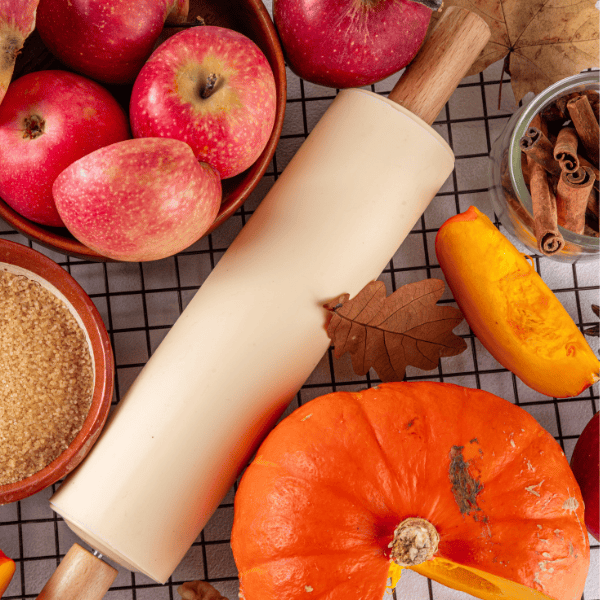Red apples, an orange pumpkin, a squash wedge, cinnamon sticks, brown sugar, and a rolling pin are arranged on a checked cloth with autumn leaves.