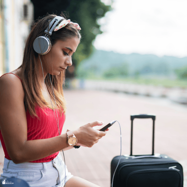 Young traveler listening to music through headphones while using a smartphone beside her suitcase at a train station.