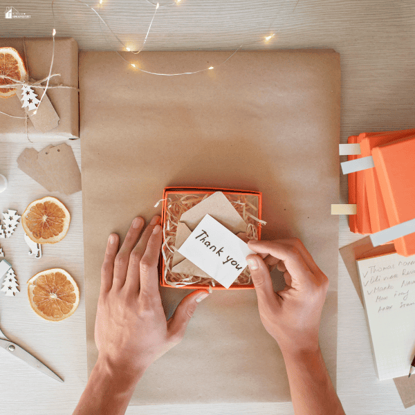 Hands creating a personalized gift box with kraft paper, decorative dried orange slices, string lights, and a thoughtful handwritten card.
