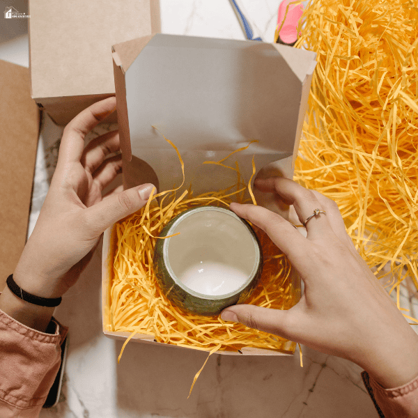 Hands arranging a ceramic cup in a personalized gift box filled with bright shredded paper for a thoughtful gift presentation.