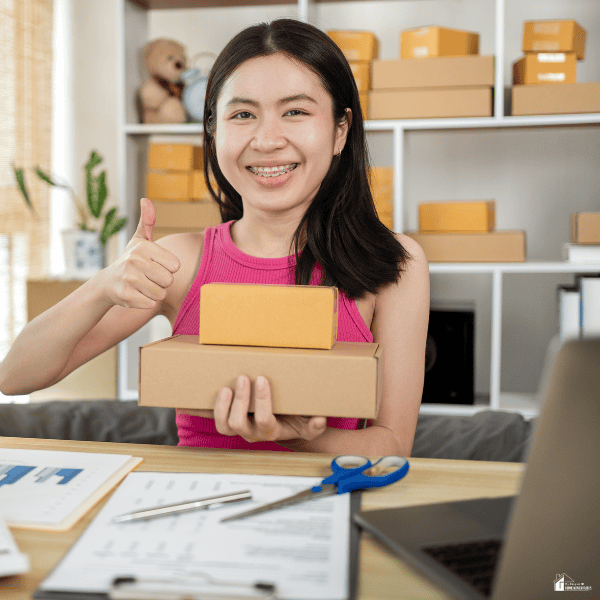Smiling home decor business owner holding packaged orders beside a laptop and shipping supplies.