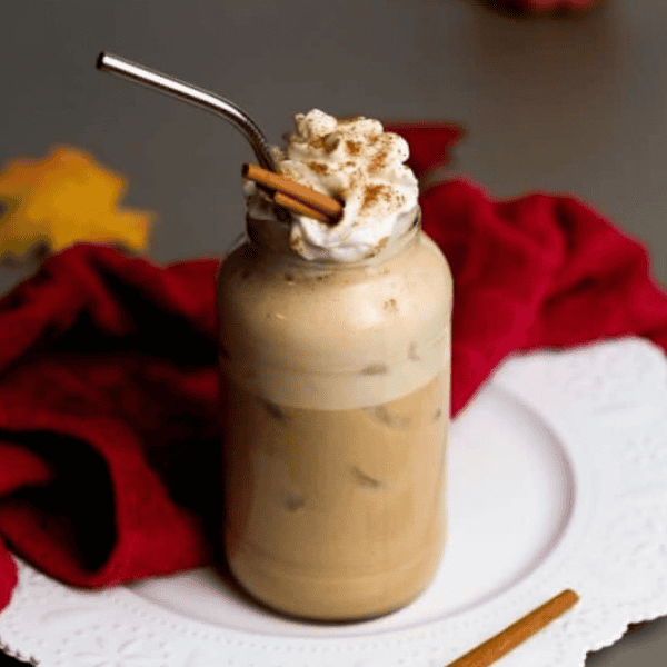 Vegan Iced Pumpkin Spice Latte: An iced pumpkin coffee drink in a glass jar is topped with whipped cream, cinnamon sticks, and a metal straw.