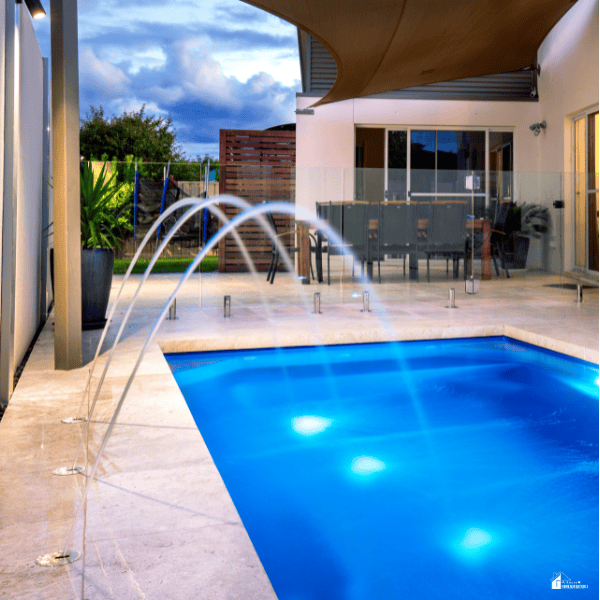 A blue pool beside a modern house has arcing water jets and clear fence panels along the deck.