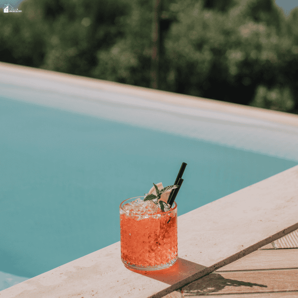 A pink drink sits on the stone edge of a pool filled with pale blue water.