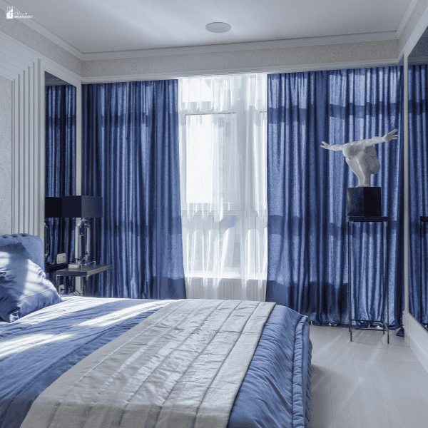Blue curtains and sheer white drapes create layered window furnishings in a bedroom.