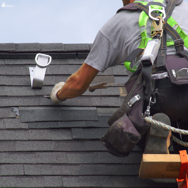 A roofer wearing a safety harness and tool belt carries out roof repair on grey asphalt shingles.
