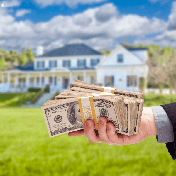 Hand holding bundles of hundred-dollar bills outside a white house, representing funds offered by cash home buyers.