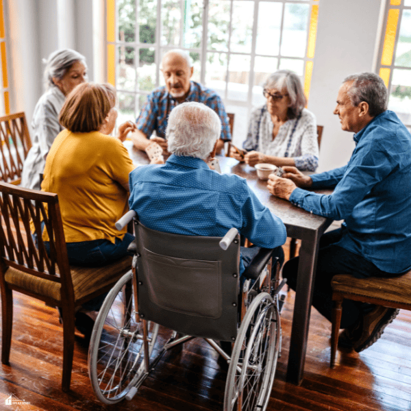 Older adults, including a wheelchair user, play cards around a table, illustrating social activities in a retirement village.