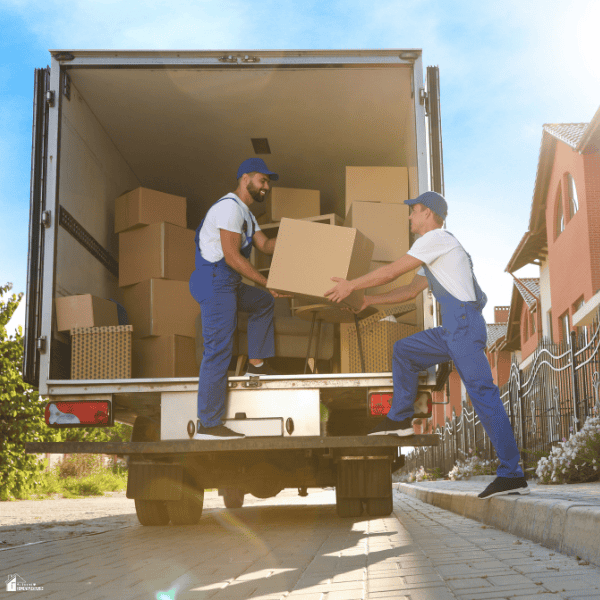 Two workers from a moving company load a cardboard box into a truck filled with household belongings.