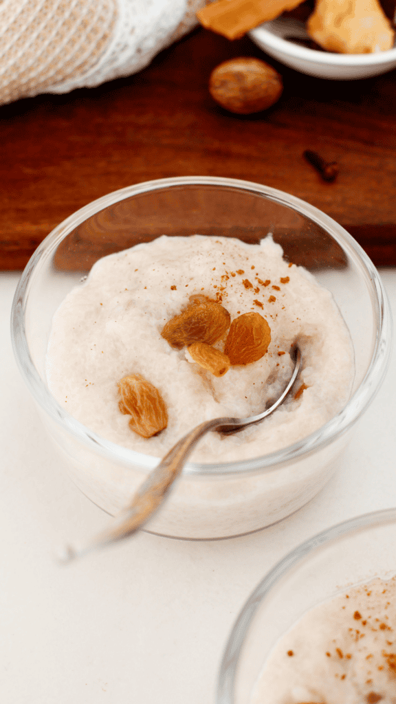 A spoonful of creamy Arroz Dulce topped with a golden raisin and cinnamon is held above a blurred glass bowl.