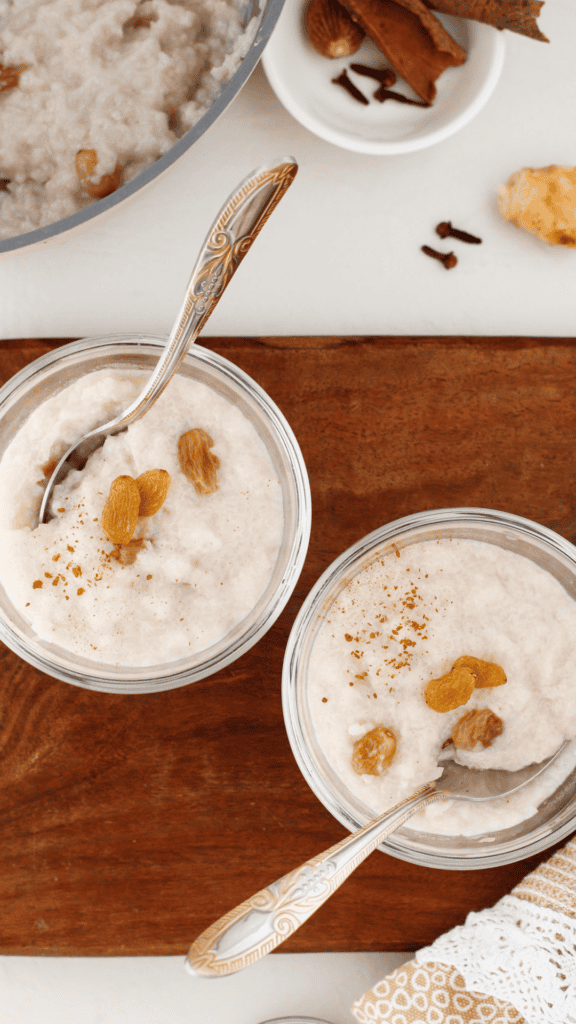 Overhead view of two glass bowls of Arroz Dulce coconut rice pudding topped with raisins and cinnamon on a wooden board with decorative silver spoons.