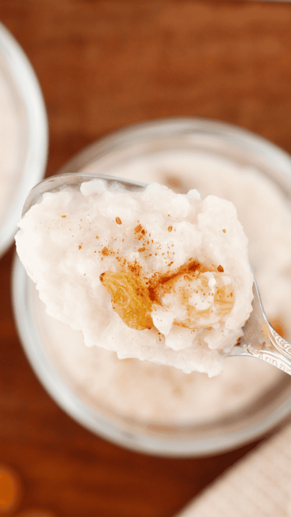A spoonful of creamy coconut rice pudding topped with a golden raisin and cinnamon is held above a blurred glass bowl.