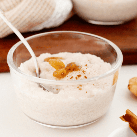 Arroz Dulce fills a clear glass bowl topped with golden raisins and cinnamon, with a silver spoon resting on the left.