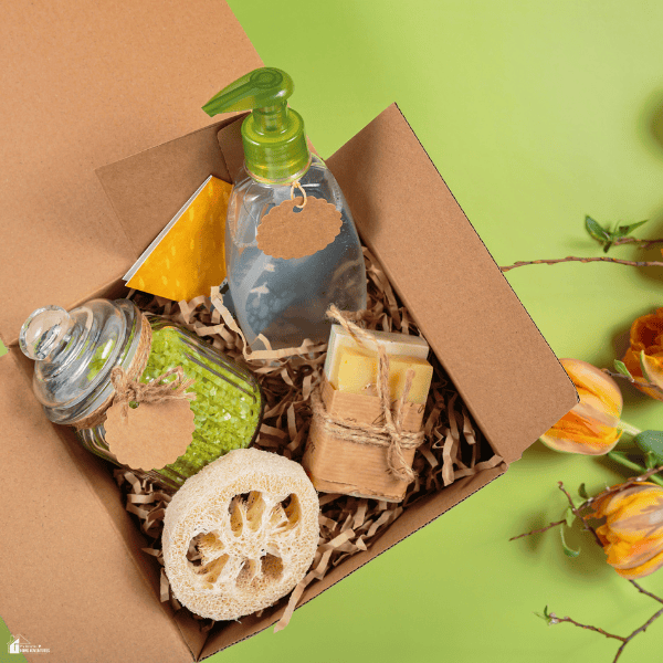 A box of hostess gifts contains bath salts, wrapped soap, a pump bottle, and a loofah beside orange flowers.