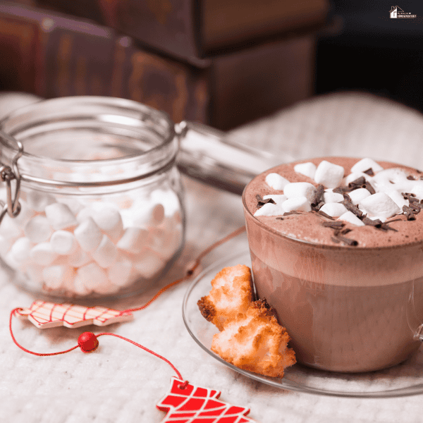 Hot chocolate topped with marshmallows sits beside cookies and a glass jar, offering inspiration for cozy hostess gifts.