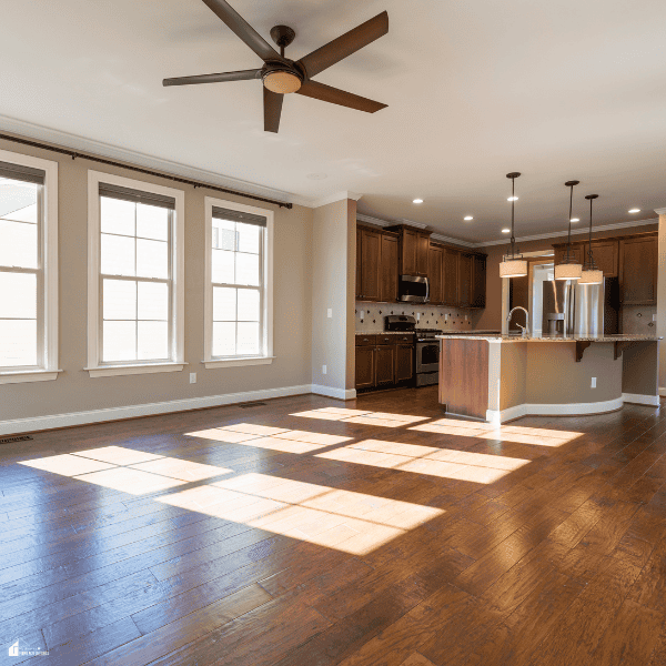 Reclaimed wood flooring in rich brown tones catches sunlight in an open living area adjoining a kitchen with dark cabinets.