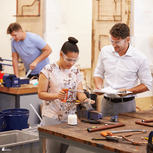 A woman demonstrates hacksaw skills while a man observes with paperwork, illustrating practical assessment relevant to an RPL training provider.