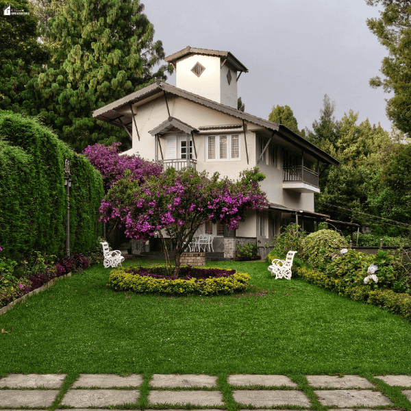 A landscaped backyard features a green lawn, flowering trees, trimmed hedges, garden beds, and white benches beside a house.