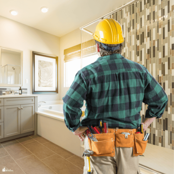 A contractor wearing a yellow hard hat and tool belt surveys a bathroom remodel beside a bathtub and mosaic-tiled shower wall.