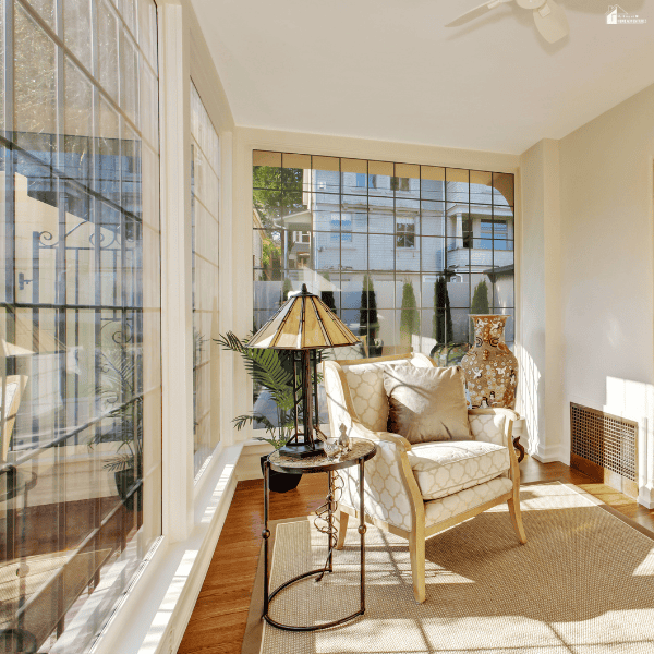 Bright sunroom with a patterned armchair, decorative lamp, potted greenery, and tall windows overlooking neighboring houses.