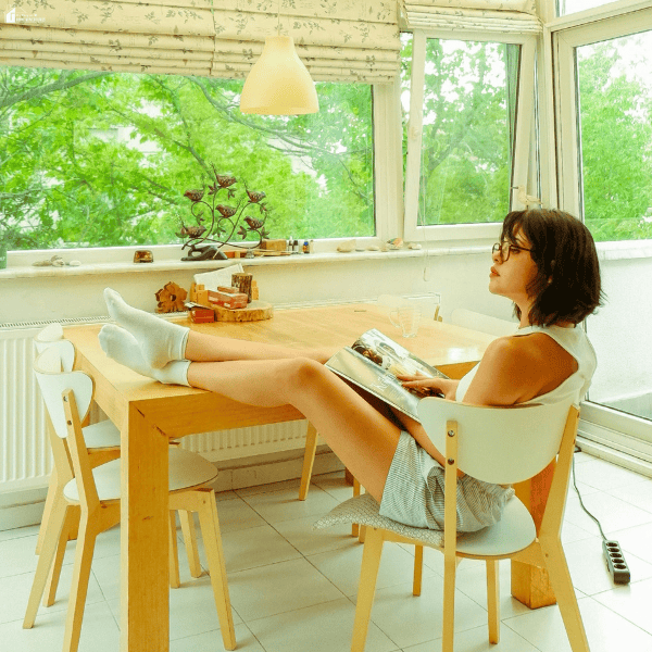 Woman reading a magazine in a sunroom with her feet resting on a wooden dining table beside windows overlooking leafy trees.