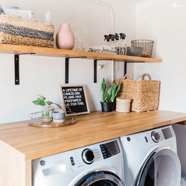 A laundry room features a wooden countertop over a washer and dryer, with open shelving, woven baskets, and cleaning supplies.