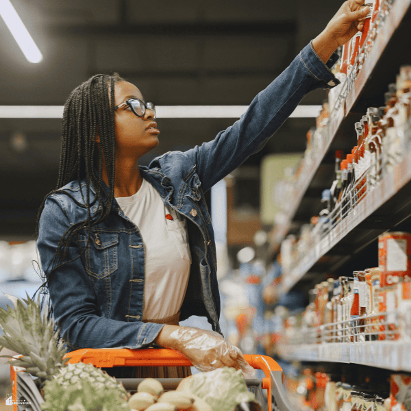 A woman selects an item from a supermarket shelf while shopping with her grocery budget in mind.