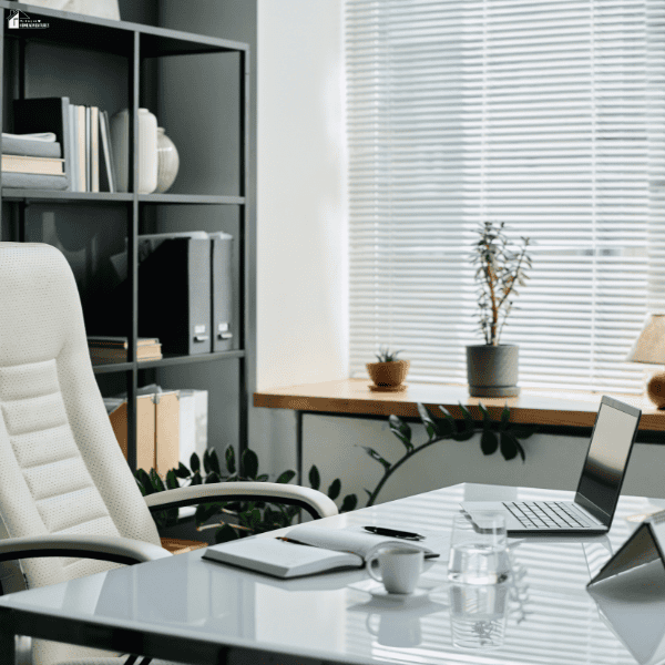A bright executive office has a glass desk, white chair, laptop, organised shelving, and plants beside window blinds.